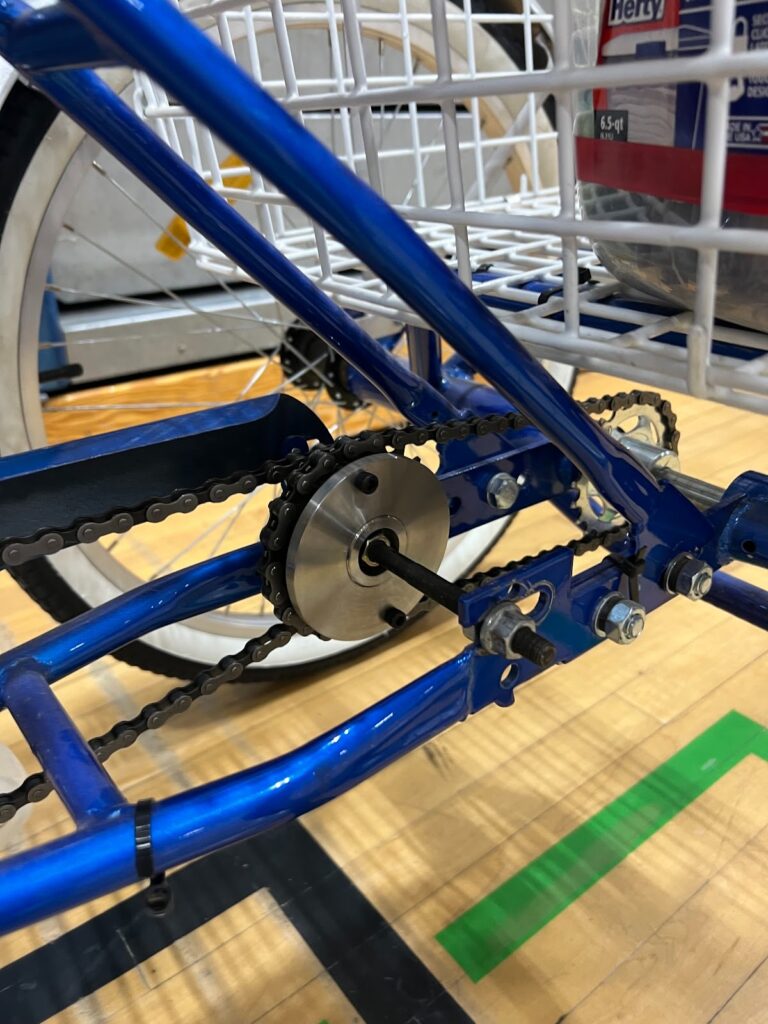 A close-up of a pedal-powered vehicle’s drivetrain, showing a metal sprocket, chain, and axle mounted on a blue frame, with part of a white basket visible in the background on a gym floor.